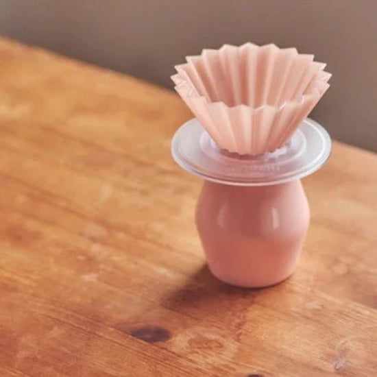 On a vintage felt wood table, a pour-over coffee set: a pink ORIGAMI AS Resin Dripper S being put on a clear ORIGAMI AS Resin Dripper Holder and a matching pink color flavor cup