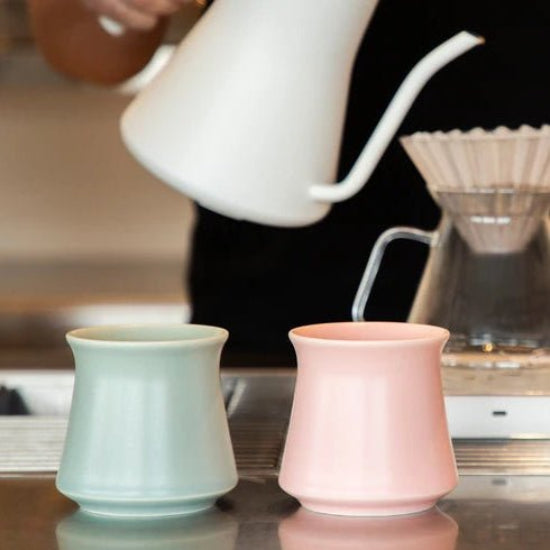 Two ceramic cups, one green and one pink, on a reflective surface with a blurred coffee-making scene in the background.