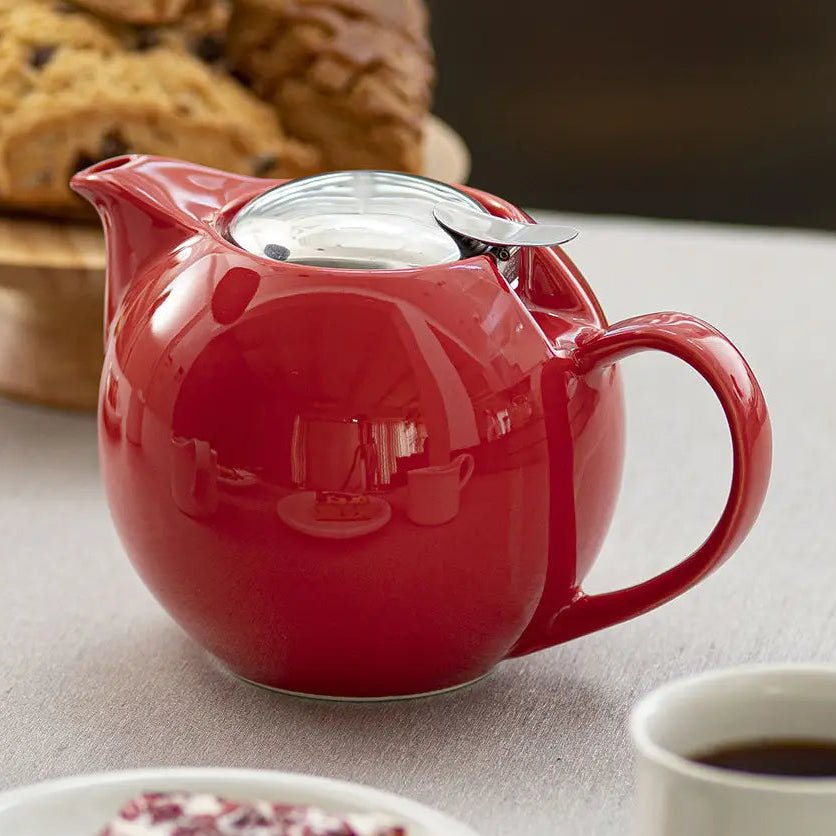 A tomato red round ceramic teapot with a silver lid on a table with bread and coffee on the table