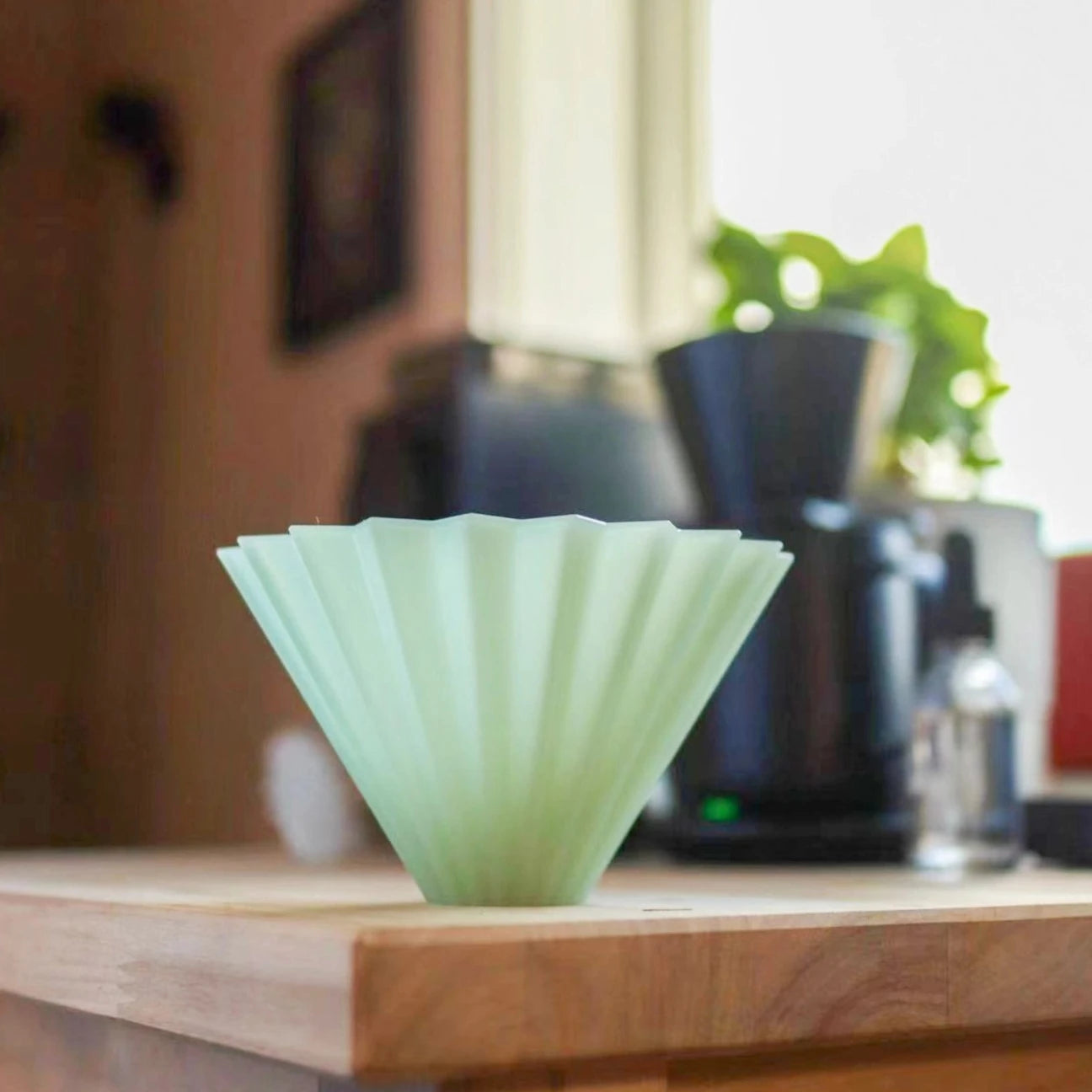 A matte green ORIGAMI Dripper Air S on a wooden table , the backdrop has some house wares and a green indoor plant 