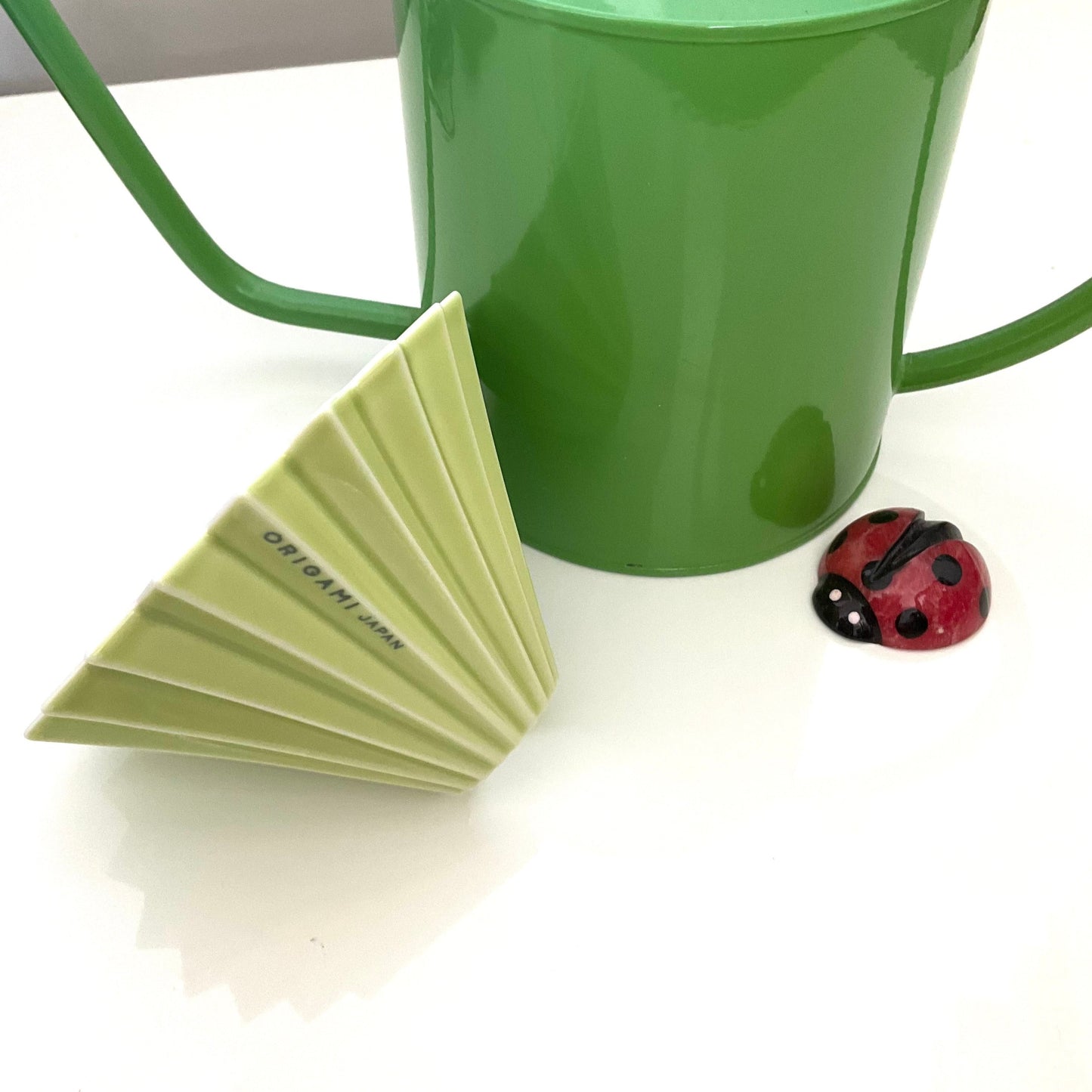 A  green RIGAMIi Coffee Dripper in front of a green flower watering pot and a small ladybug figure