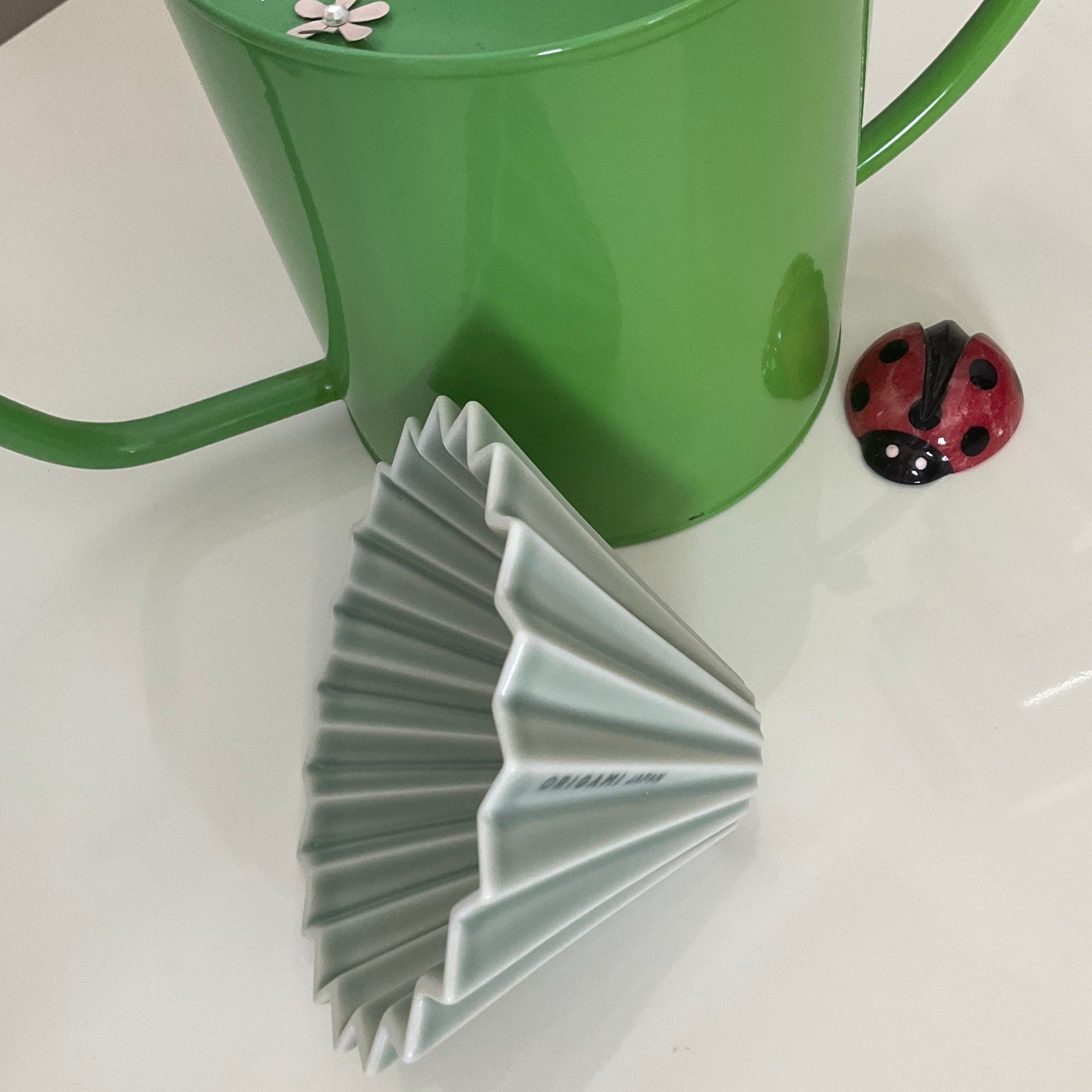 Front is a matte green ORIGAMI Coffee Dripper, Medium, at the back is a green watering jar, and a small ladybug figure at side