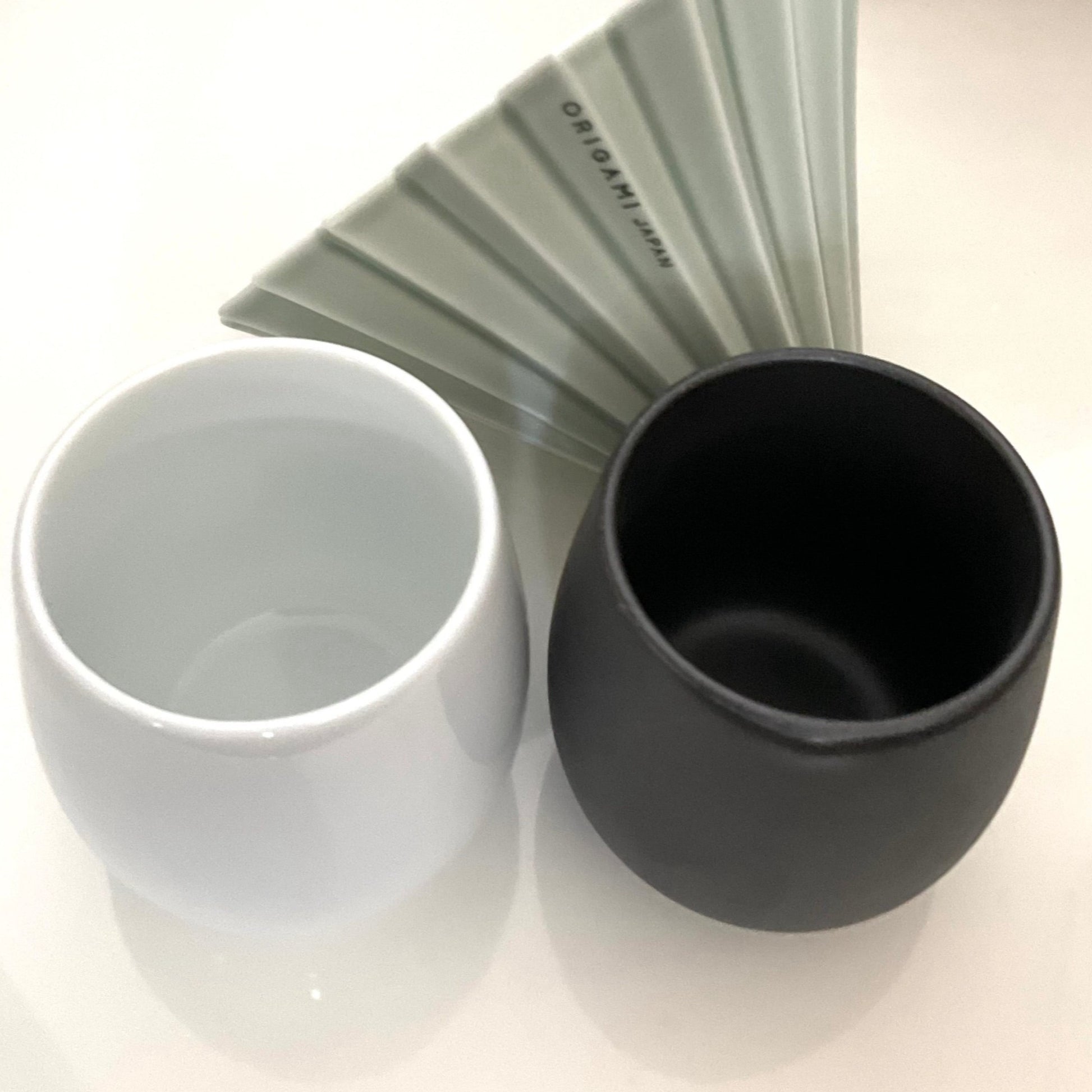 A white and a matte black ORIGAMI Barrel Flavor Cups put together and a matte green ORIGAMI Coffee Dripper at the back