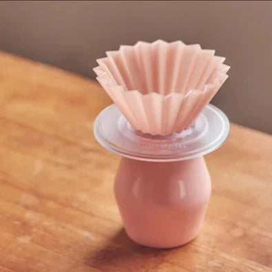 On a wood table, there is a matte pink ORIGAMI Dripper Air S (made of AS resin) being put on a clear plastic ORIGAMI dripper holder and on a pink flavor cup