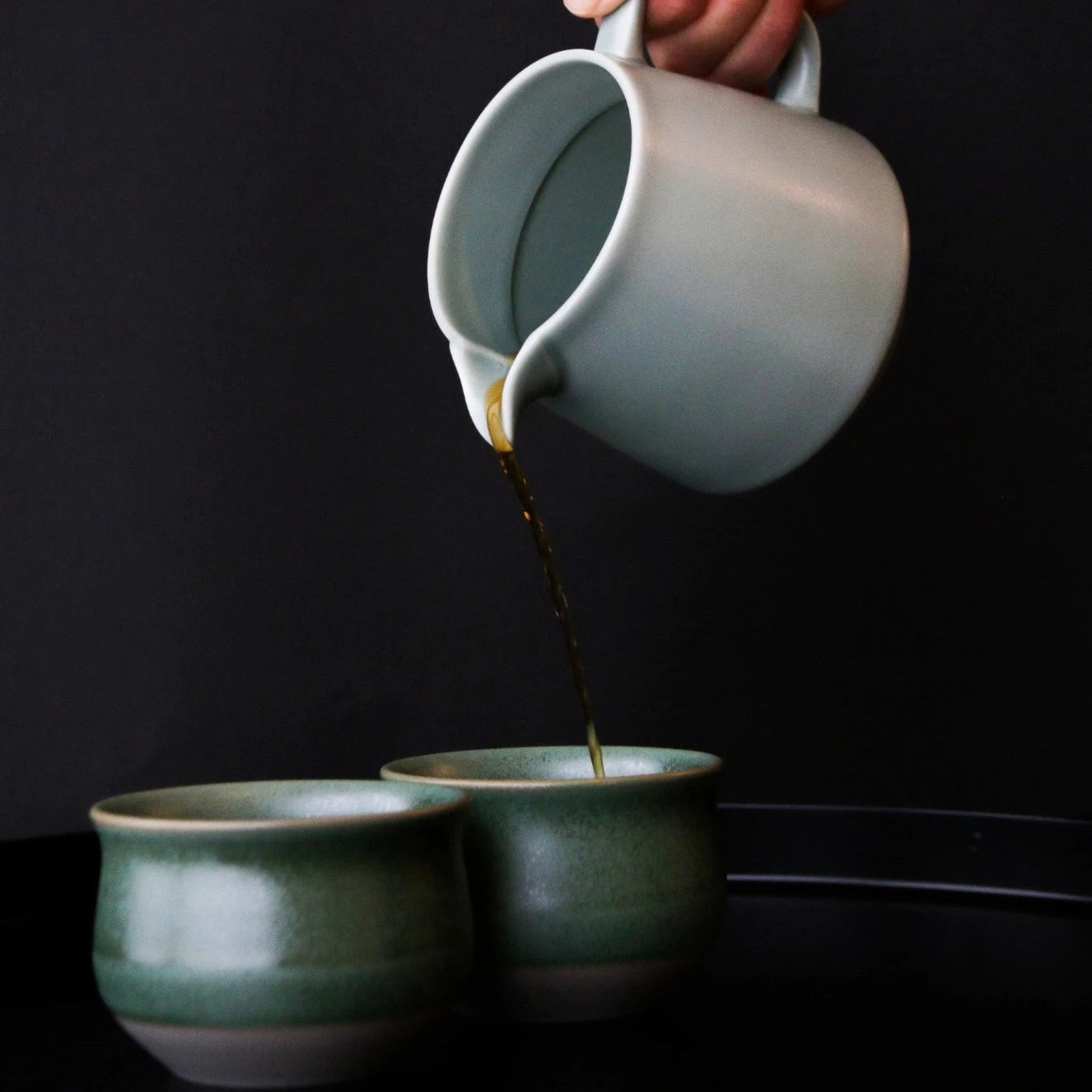 Pouring coffee from a matte gray ORIGAMI Ceramic Coffee server into two pine green ORIGAMI Pinot Flavor Bowls