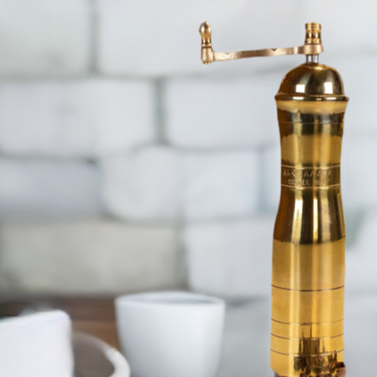 Athena 8.7" Brass Manual Coffee Grinder on a table with white latte cup next to it and white stone wall backdrop