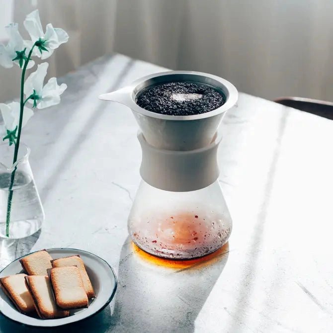 Coffee maker with ground coffee on top half of the device and a plate of cookies and a vase with flowers next to it