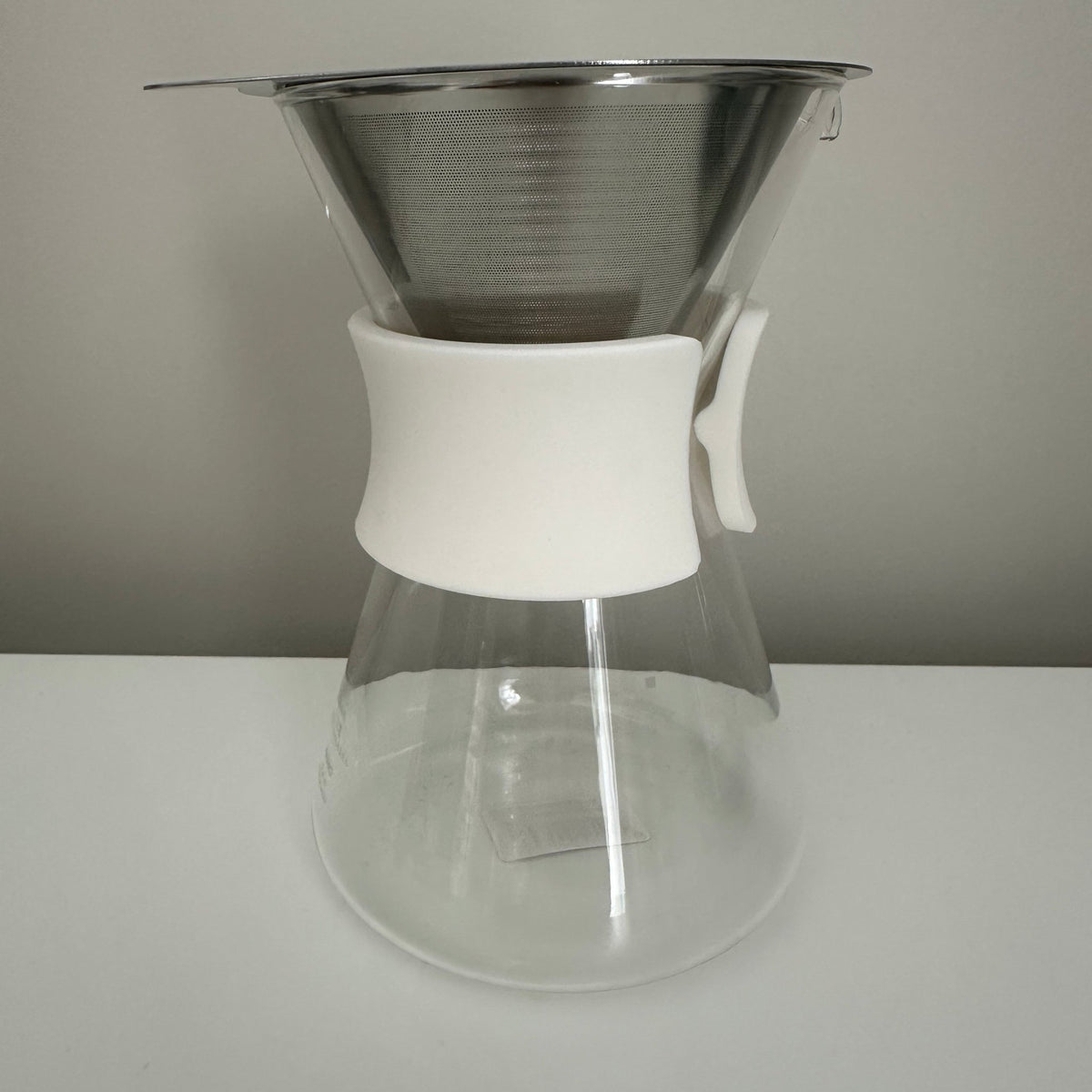 A manual pour-over coffee maker with a clear glass carafe, a white plastic handle, and a stainless steel mesh filter cone on top, set against a neutral gray background.