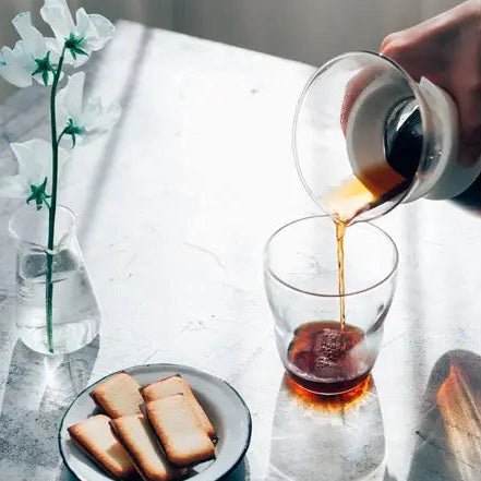 A person pouring coffee into a glass on a marble surface with a vase and cookies.