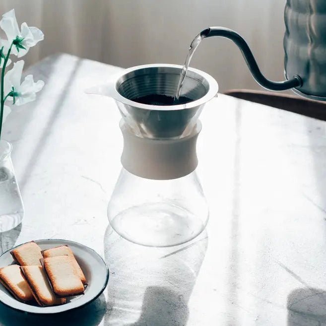 Water being poured into a glass carafe on a light surface with a plate of cookies and a vase of flowers.