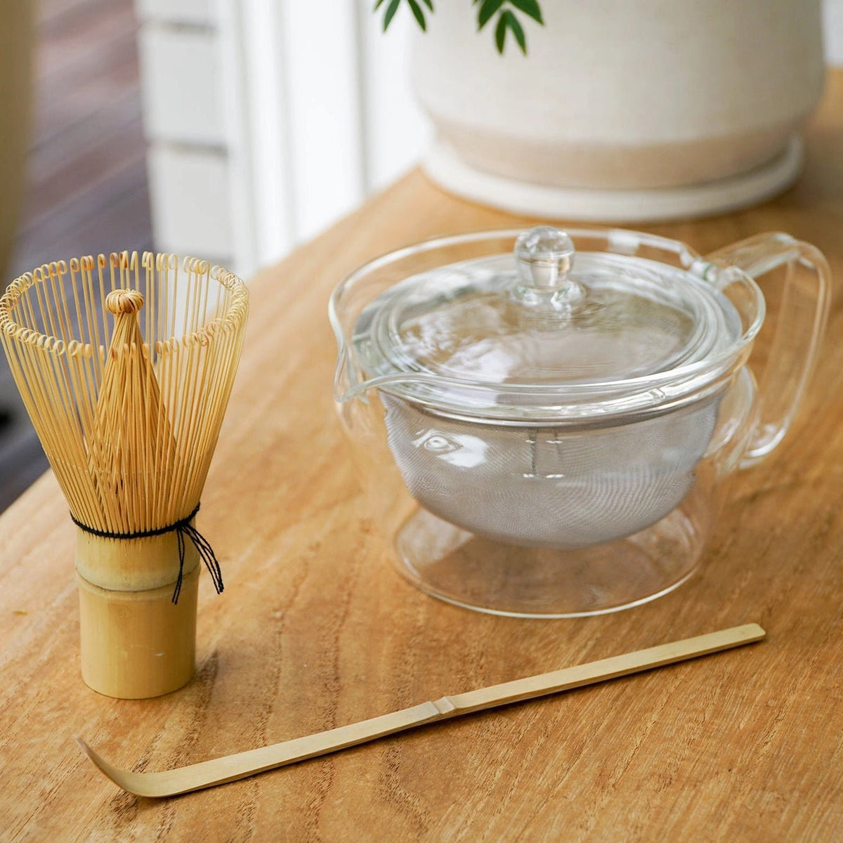 A glass teapot with a lid and strainer sits on a wooden table alongside a bamboo whisk and bamboo scoop, with a blurred potted plant in the background.
