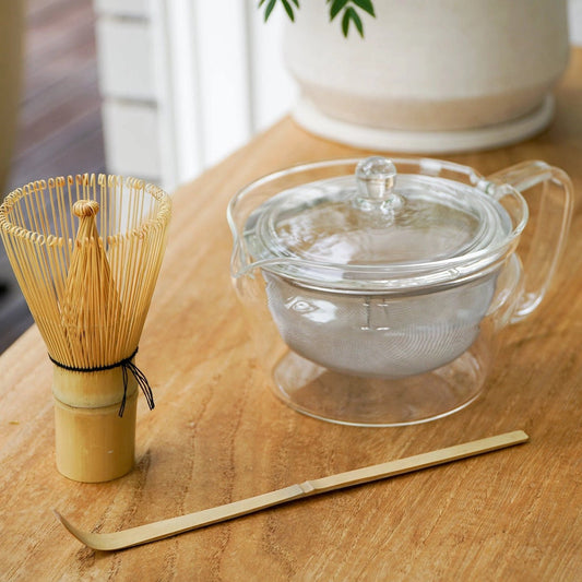 A glass teapot with a lid and strainer sits on a wooden table alongside a bamboo whisk and bamboo scoop, with a blurred potted plant in the background.
