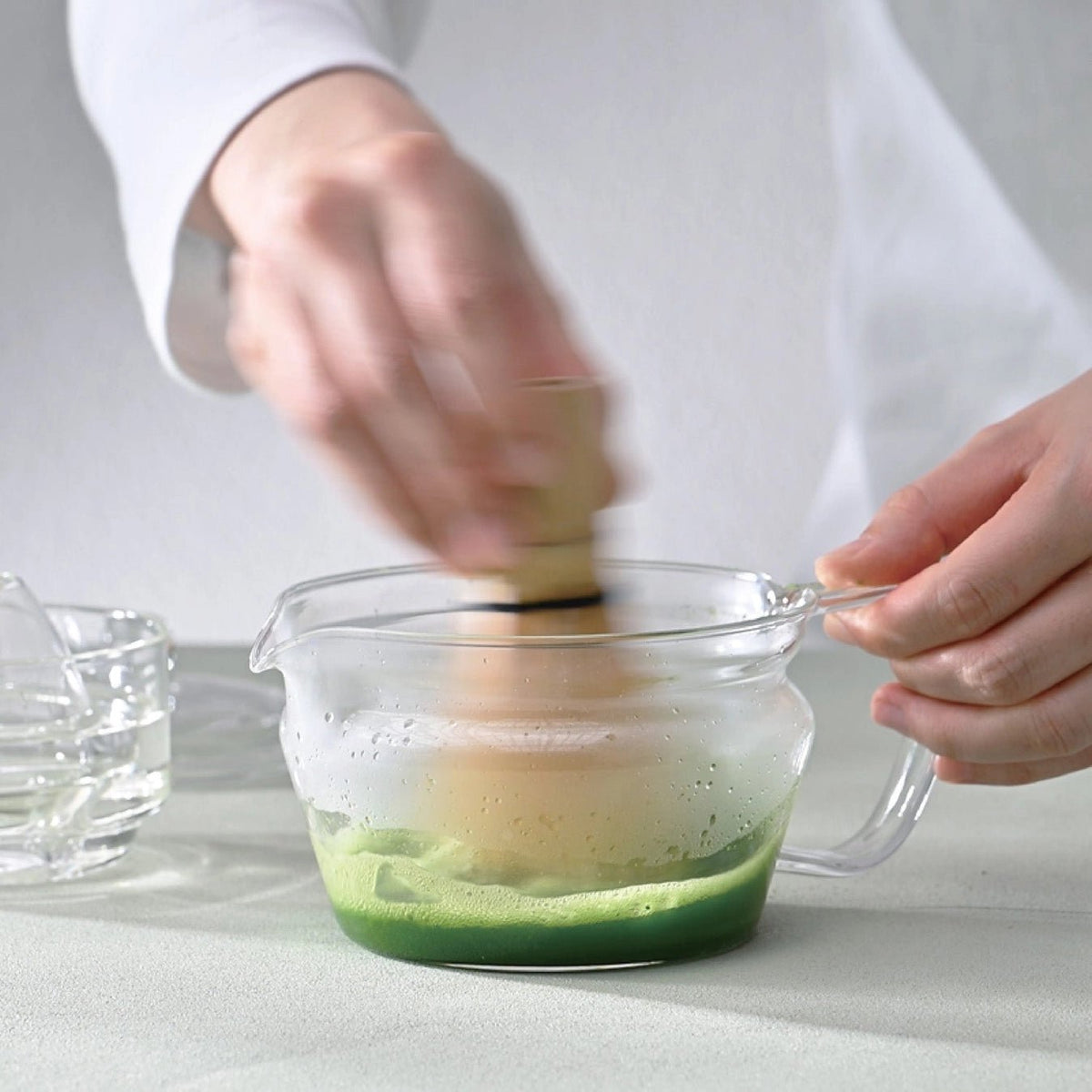 A person in a white uniform holds a bamboo whisk over a frosted glass pitcher containing green matcha tea, with a small glass bowl nearby on a light gray surface.
