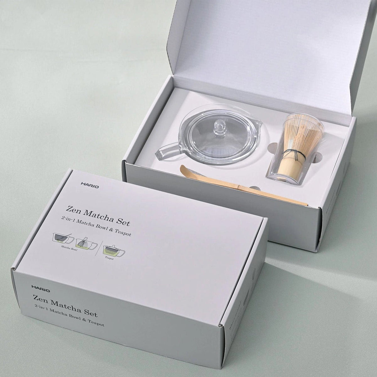 A HARIO Zen Matcha Set, shown with its white packaging box in the foreground and the open box displaying the glass teapot, bamboo whisk, and wooden scoop on a light gray surface.