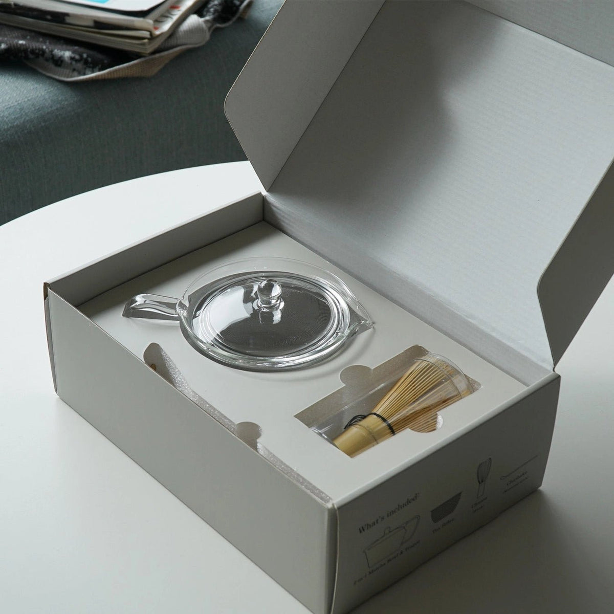 A clear glass teapot with a lid sits inside an open white gift box, alongside a bamboo whisk and a small bamboo scoop. The box is placed on a white table with a gray upholstered chair in the background.