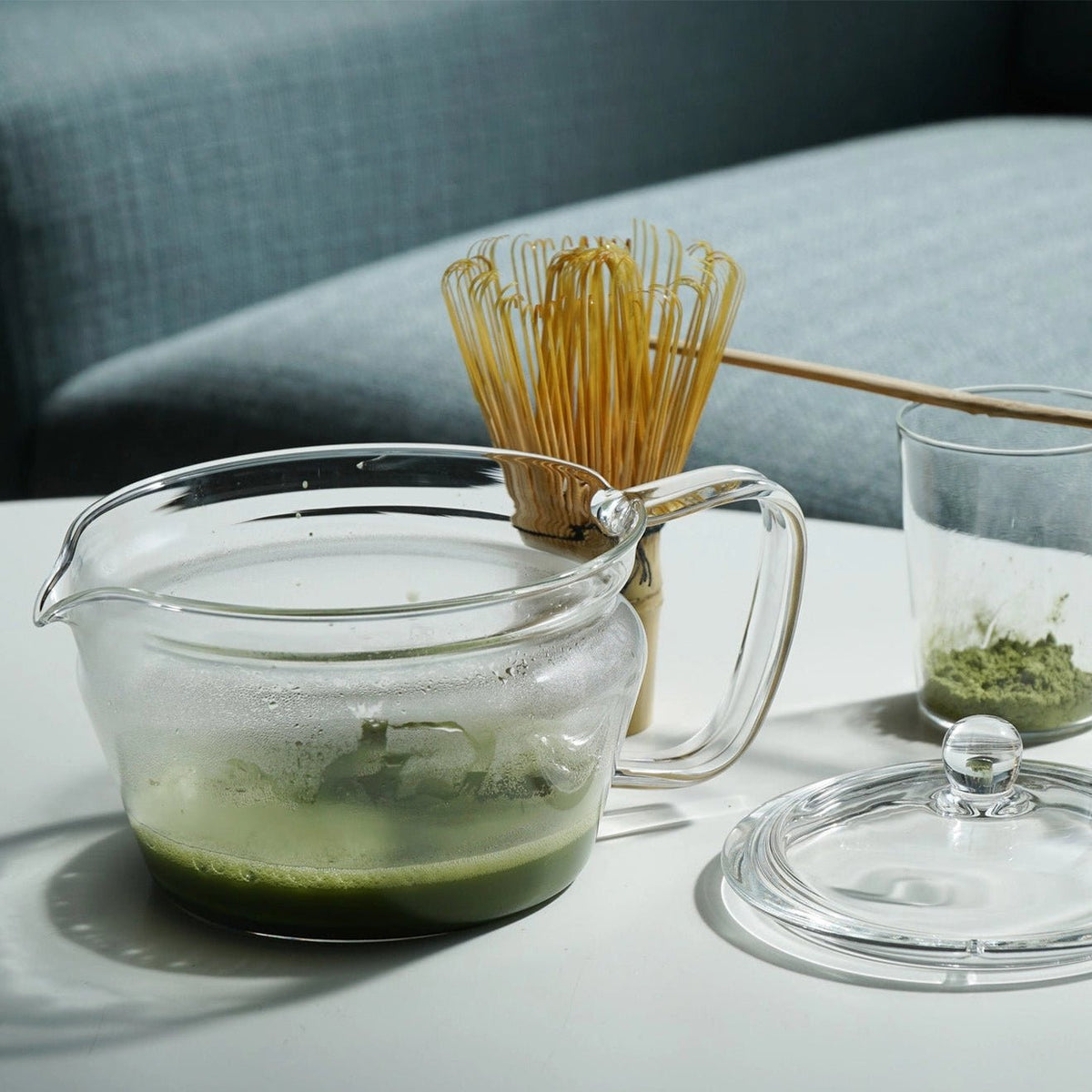 A clear glass matcha whisking pitcher filled with green tea sits on a white table, with a bamboo whisk and glass cup of matcha powder nearby against a blue-gray upholstered chair background.