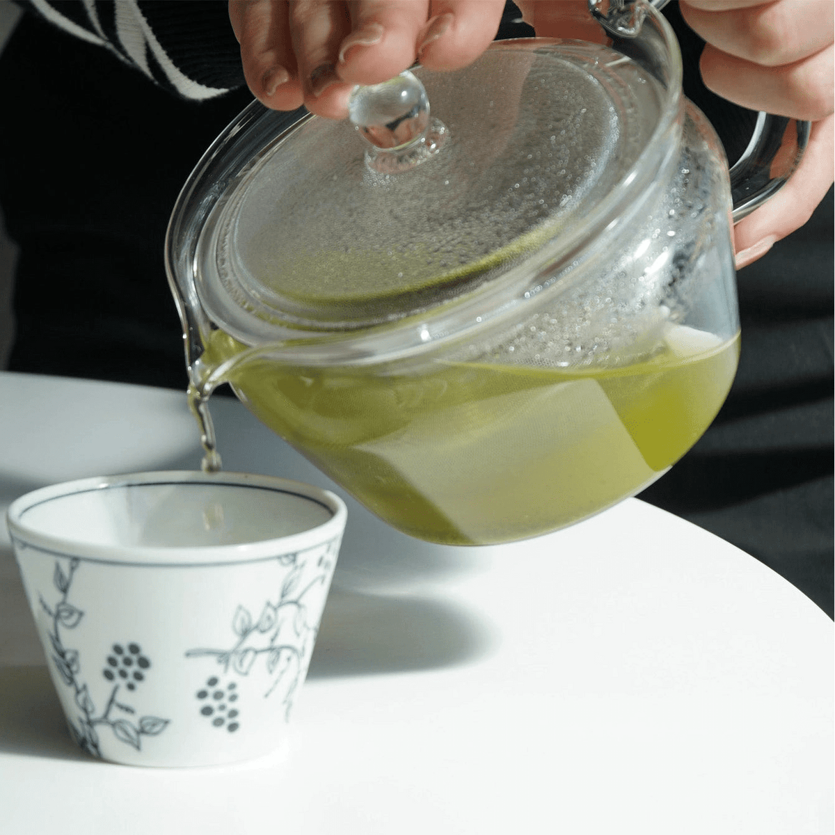 A clear glass teapot with a lid is being held by hands and used to pour green tea into a small white ceramic cup with blue floral patterns, set on a white surface.