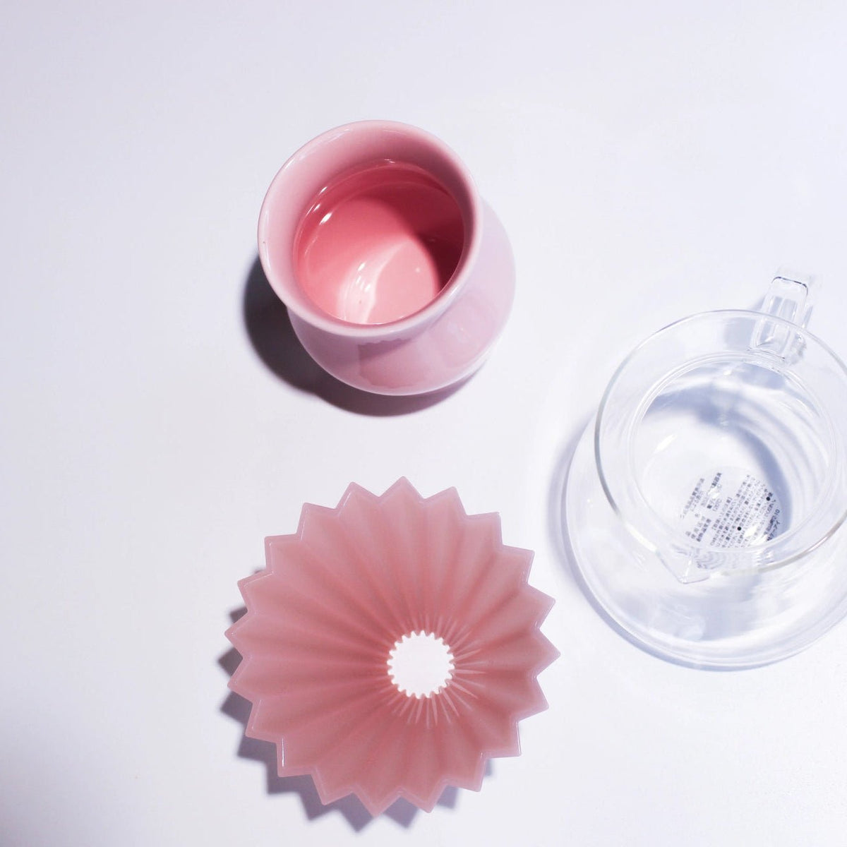 A pink plastic coffee filter cone and a matching pink ceramic cup are arranged on a white surface next to a clear glass teacup with a clear silicone infuser.